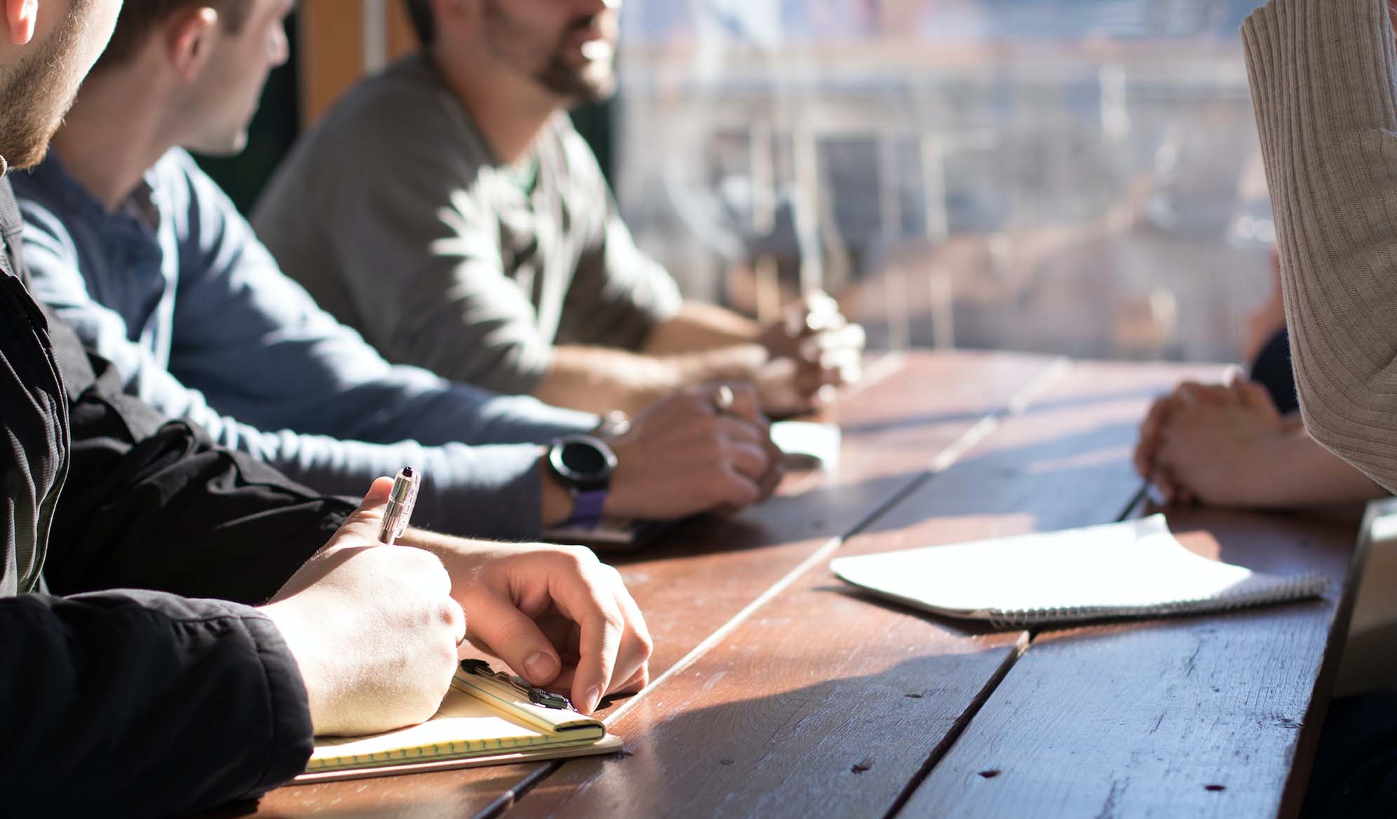 Four people seated at a table talking, one is writing in a notebook.