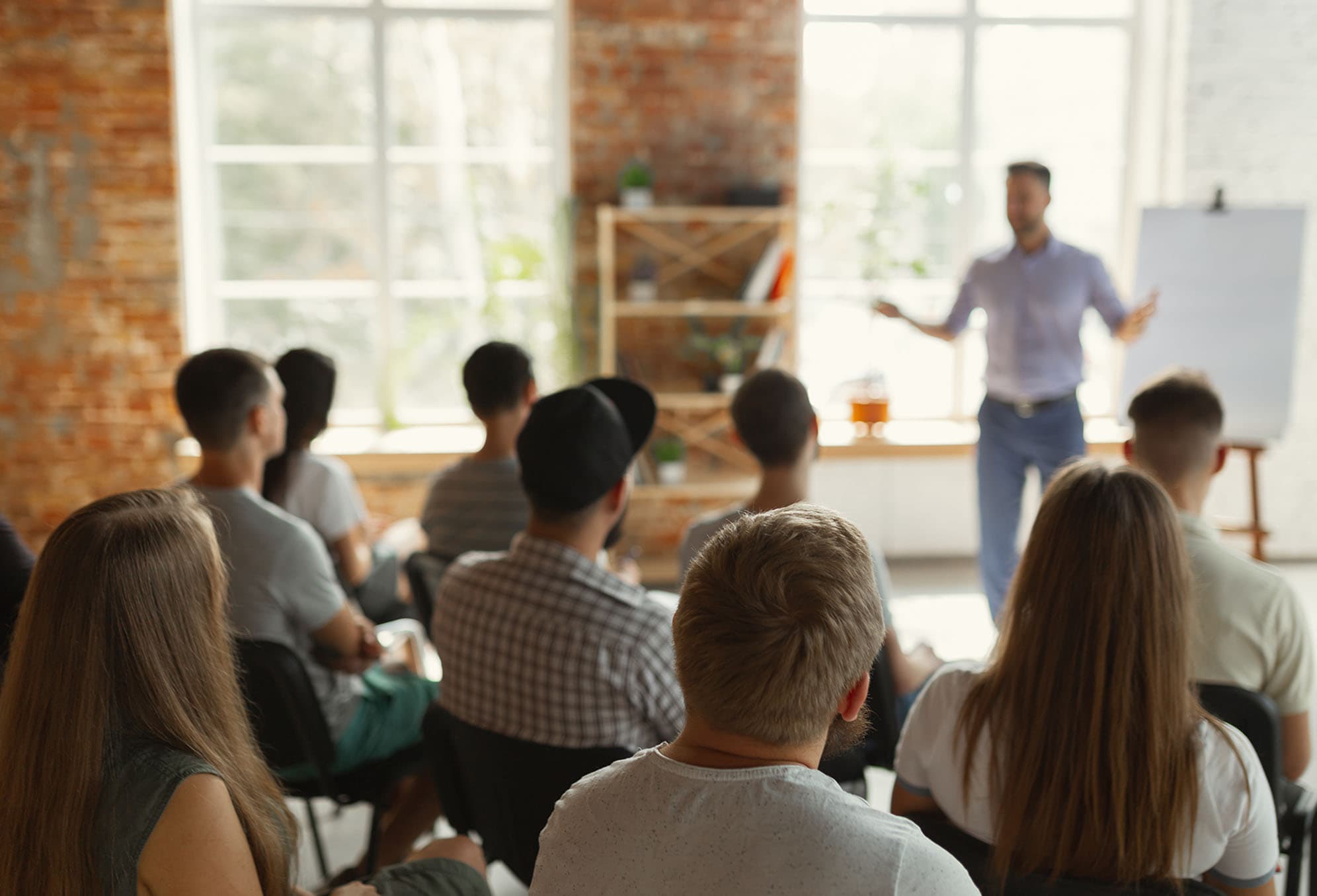 A classroom setting with large windows, and rows of people from the back who are  listening to a man standing in front of a white board and easel talking.