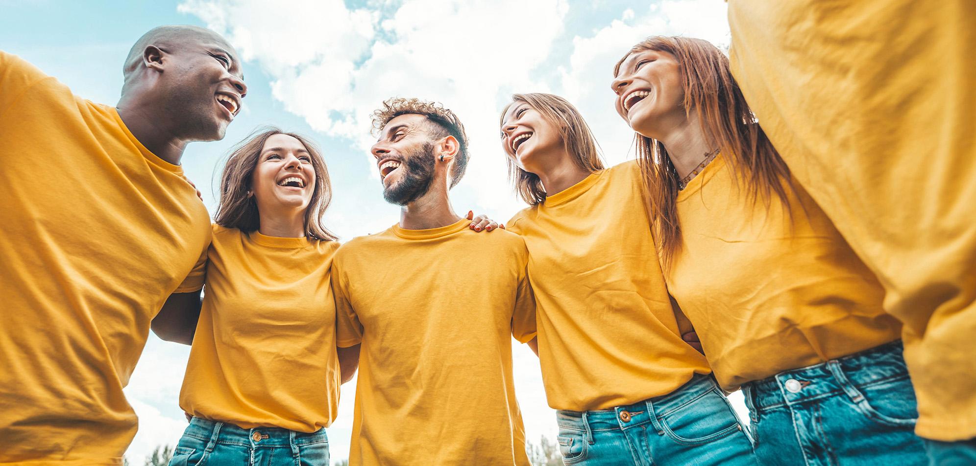 A diverse group of laughing people wearing yellow shirts and standing in a circle with their arms around one another.