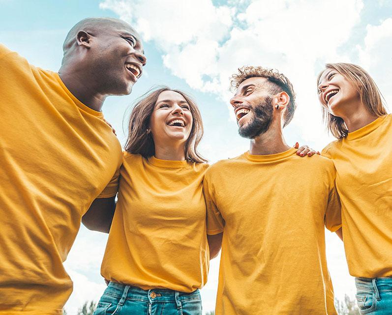 A diverse group of laughing people wearing yellow shirts and standing in a circle with their arms around one another.